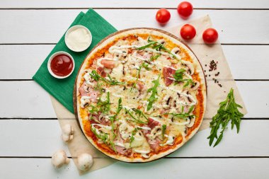 Salamlı, rukkola ve mantarlı taze pişmiş pizza. Ahşap zemin üzerinde domates, sos ve otlarla servis ediliyor. Yemek teslimatı konsepti. Restoran menüsü. Üst görünüm