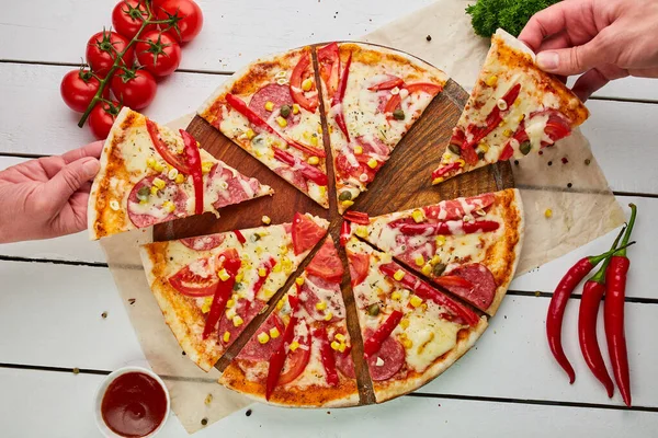 The human hand takes Freshly baked tasty pepperoni pizza with salami, mozzarella cheese, corn and pepper served on wooden background with tomatoes, sauce and herbs. Food delivery concept. Restaurant menu