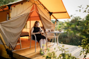 Mutlu Kadın Serbest Çalışan, güneşli bir günde rahat bir glamping çadırında dinlenirken telefonla içki içiyor. Yaz tatili ve tatil için lüks kamp çadırı. Yaşam tarzı kavramı