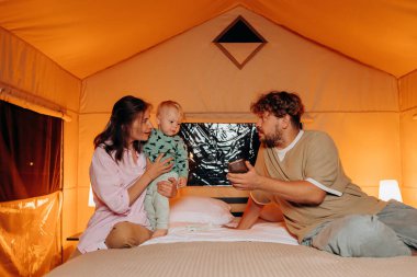 Happy family with lovely baby playing and spend time together in cozy glamping on summer evening while lying on bed. Luxury camping tent for outdoor recreation and recreation