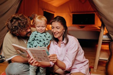 Güzel bir bebeği olan mutlu bir aile yaz akşamları glamping 'de birlikte kitap okuyor. Açık hava eğlencesi ve dinlenme için lüks kamp çadırı. Yaşam tarzı kavramı