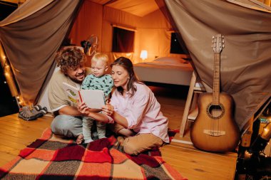 Güzel bir bebeği olan mutlu bir aile yaz akşamları glamping 'de birlikte kitap okuyor. Açık hava eğlencesi ve dinlenme için lüks kamp çadırı. Yaşam tarzı kavramı