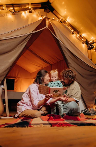 Güzel bir bebeği olan mutlu bir aile yaz akşamları glamping 'de birlikte kitap okuyor. Açık hava eğlencesi ve dinlenme için lüks kamp çadırı. Yaşam tarzı kavramı