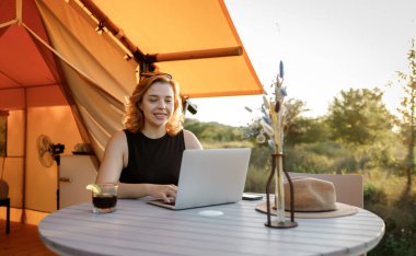 Smiling Woman freelancer using a laptop on a cozy glamping tent in a sunny day. Luxury camping tent for outdoor summer holiday and vacation. Lifestyle concept