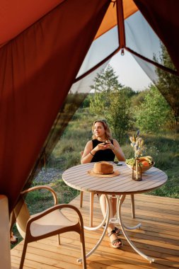 Mutlu Kadın Serbest Çalışan, güneşli bir günde rahat bir glamping çadırında dinlenirken telefonla içki içiyor. Yaz tatili ve tatil için lüks kamp çadırı. Yaşam tarzı kavramı