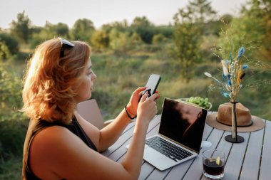 Güler yüzlü kadın, güneşli bir günde rahat bir çadırda dizüstü bilgisayarla çalışırken telefonu kullanıyor. Yaz tatili ve tatil için lüks kamp çadırı. Yaşam tarzı kavramı