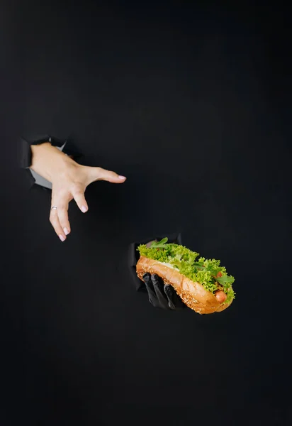 Hand holding hot dog through torn holes in black paper background