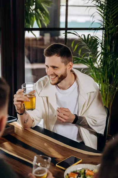 Barda ya da barda arkadaşlarıyla eğlenirken bira içen ve gülen genç adam.