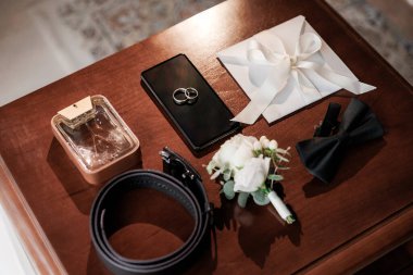 Damatların düğün gereksinimleri masada, telefondaki alyanslar, papyon, yaka çiçeği, parfüm, kemer ve kurdeleli zarf. Düğün hazırlıkları, damat aksesuarları için ideal.