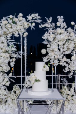 Çiçek aksanlı, minimalist üç kademeli beyaz bir pasta ile zarif bir düğün hazırlığı. Gece gökyüzünün altında, yemyeşil beyaz çiçeklerle çevrili. Lüks etkinlikler ve zarif kutlamalar.