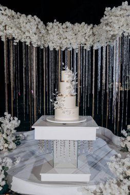 Çiçek aksanlı, minimalist üç kademeli beyaz bir pasta ile zarif bir düğün hazırlığı. Gece gökyüzünün altında, yemyeşil beyaz çiçeklerle çevrili. Lüks etkinlikler ve zarif kutlamalar.