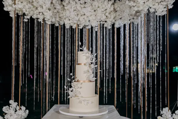 stock image Elegant wedding setup with a minimalist three-tier white cake with fresh floral accents, surrounded by lush white floral arrangements under the night sky. Luxury events, and elegant celebrations