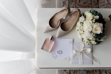 Gelinlerin düğün gereksinimleri: Topuklar, buket, yüzükler, parfüm ve davetiye beyaz bir masada, zarif bir hazırlık. Yüksek kalite fotoğraf