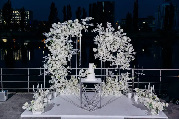 stock image Elegant wedding setup with a minimalist three-tier white cake with fresh floral accents, surrounded by lush white floral arrangements under the night sky. Luxury events, and elegant celebrations