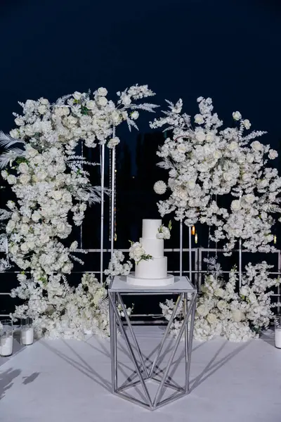 stock image Elegant wedding setup with a minimalist three-tier white cake with fresh floral accents, surrounded by lush white floral arrangements under the night sky. Luxury events, and elegant celebrations