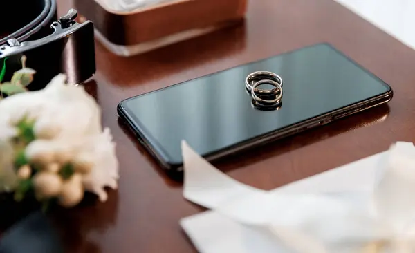 stock image Grooms wedding essentials on table, featuring wedding rings on phone, bow tie, boutonniere, perfume, belt, and ribbon-tied envelope. Ideal for wedding preparations, grooms accessories