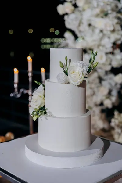 Stock image Elegant wedding setup with a minimalist three-tier white cake with fresh floral accents, surrounded by lush white floral arrangements under the night sky. Luxury events, and elegant celebrations