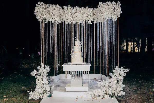 stock image Elegant wedding setup with a minimalist three-tier white cake with fresh floral accents, surrounded by lush white floral arrangements under the night sky. Luxury events, and elegant celebrations