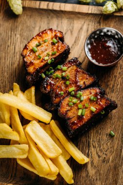 Close-up of barbecue ribs with crispy fries on a wooden board, served with a side of dipping sauce. High quality photo clipart