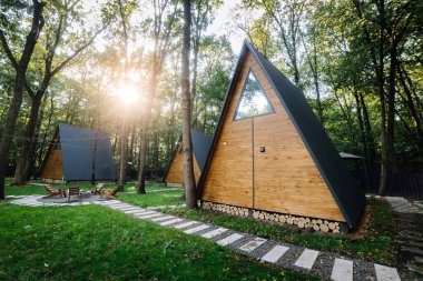 Cozy wooden A-frame cabins in a forest with morning sunlight and outdoor seating area. High quality photo clipart