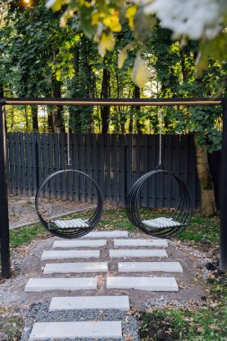 Çevresi yemyeşil ağaçlar ve güneş ışığıyla çevrili, rahat bir bahçe ortamında modern salıncaklar. Yüksek kalite fotoğraf