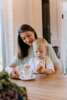 Anne ve çocuk, mutfakta kahve ve kruvasanla kahvaltı edip huzurlu bir anı paylaşıyorlar. Yüksek kalite fotoğraf