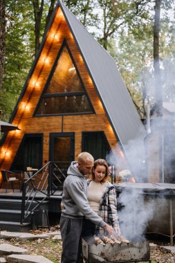 Couple grilling skewers together, enjoying barbecue in forest setting with firewood and nature. High quality photo clipart