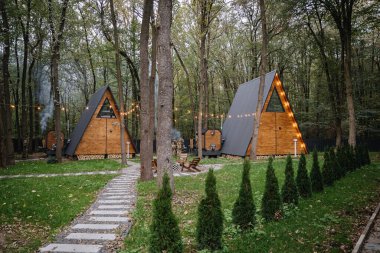 Cozy wooden A-frame cabins in a forest with morning sunlight and outdoor seating area. High quality photo clipart