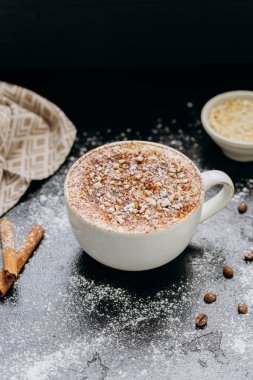 Cup of cappuccino with cinnamon, nuts, and powdered sugar on dark rustic surface with coffee beans. High quality photo clipart