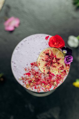 Martini bardağındaki zarif mor kokteyl, kurutulmuş limon, taç yaprakları ve yenilebilir çiçeklerle süslenmiş. Yüksek kalite fotoğraf