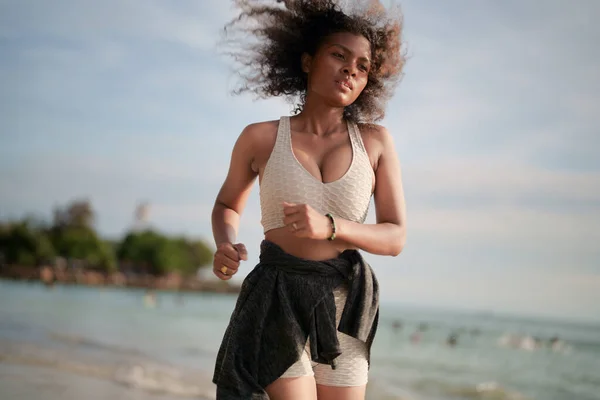 African woman in sport bra is running on beach for outdoor workout. Portrait sexy Asian African lady preparing herself for fitness at beach.