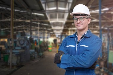 El işçisinin portresi mavi çalışma odası elbisesi ve güvenlik kaskıyla ağır sanayi fabrikası için gerekli ekipman ve ön makinede duruyor. Üretim için çelik fabrikası.