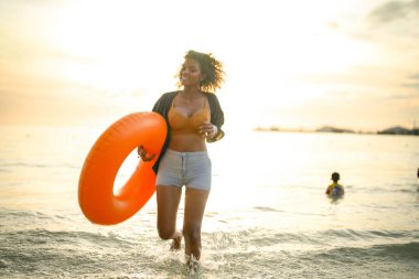 Seksi Asyalı Afrikalı kadın sahilde durup dinleniyor. Deniz ve sahilde yaz mevsimi. Tropik plajda sarı yüzme ringi oynayan kadın..