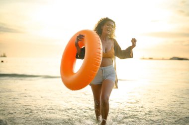 Seksi Asyalı Afrikalı kadın sahilde durup dinleniyor. Deniz ve sahilde yaz mevsimi. Tropik plajda sarı yüzme ringi oynayan kadın..