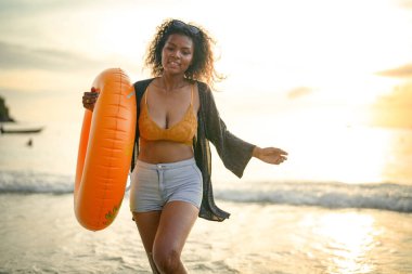 Seksi Asyalı Afrikalı kadın sahilde durup dinleniyor. Deniz ve sahilde yaz mevsimi. Tropik plajda sarı yüzme ringi oynayan kadın..