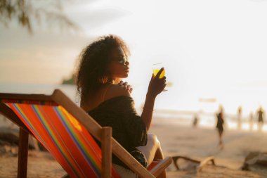 Afrikalı bir kadın tropik plajda bankta otururken kokteyl suyu içiyor. Sarı bikini giyen ve doğanın güzelliğini ürperten genç bir gezgin. Seyahat ve seyahat kavramı. 