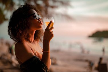 Afrikalı bir kadın tropik plajda bankta otururken kokteyl suyu içiyor. Sarı bikini giyen ve doğanın güzelliğini ürperten genç bir gezgin. Seyahat ve seyahat kavramı. 