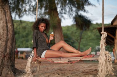 Afrikalı bir kadın tropik plajda salıncakta sallanırken kokteyl suyu içiyor. Sarı bikini giyen ve doğanın güzelliğini ürperten genç bir gezgin. Seyahat ve seyahat kavramı. 