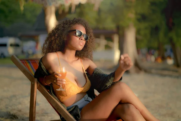 stock image African woman drinking cocktail juice when sitting on bench beach at tropical beach. Young traveler wearing yellow bikini and chilling out the beauty of the Nature. Wanderlust and travel concept. 