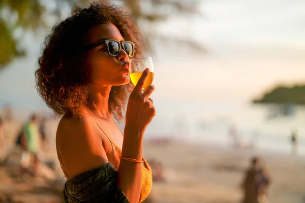 stock image African woman drinking cocktail juice when sitting on bench beach at tropical beach. Young traveler wearing yellow bikini and chilling out the beauty of the Nature. Wanderlust and travel concept. 