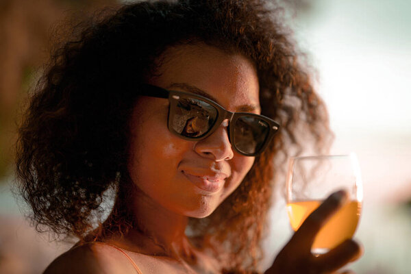 African woman drinking cocktail juice when sitting on bench beach at tropical beach. Young traveler wearing yellow bikini and chilling out the beauty of the Nature. Wanderlust and travel concept. 