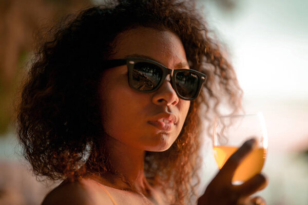 African woman drinking cocktail juice when sitting on bench beach at tropical beach. Young traveler wearing yellow bikini and chilling out the beauty of the Nature. Wanderlust and travel concept. 
