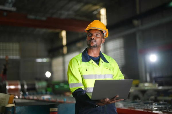 工場のエンジニアアフリカの男はラップトップで自信を持って立っている 労働者は工場の重機で働いている 機械設備プラント技術で — ストック写真