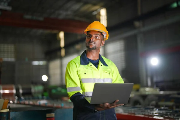 Factory engineer African man standing confidence with laptop. Worker works at heavy machine at industry factory. with machinery equipment plant technology.