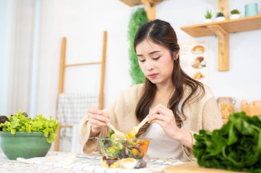 Genç Asyalı kadın salata sebzesiyle sağlıklı yiyecekler yiyor. Güzel bir iç mutfakta kilerde oturan bir kadın. Yerel ürünlerden ve malzemelerden alınan temiz diyet gıdaları taze pazardır..