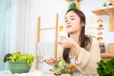 Genç Asyalı kadın salata sebzesiyle sağlıklı yiyecekler yiyor. Güzel bir iç mutfakta kilerde oturan bir kadın. Yerel ürünlerden ve malzemelerden alınan temiz diyet gıdaları taze pazardır..