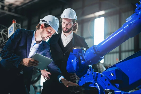 Business engineers meet and check control heavy machine robot arm. Diverse Team of Industrial Robotics Engineers Gathered Around machine. Professional Machinery Operator Use Industrial Digital Tablet.