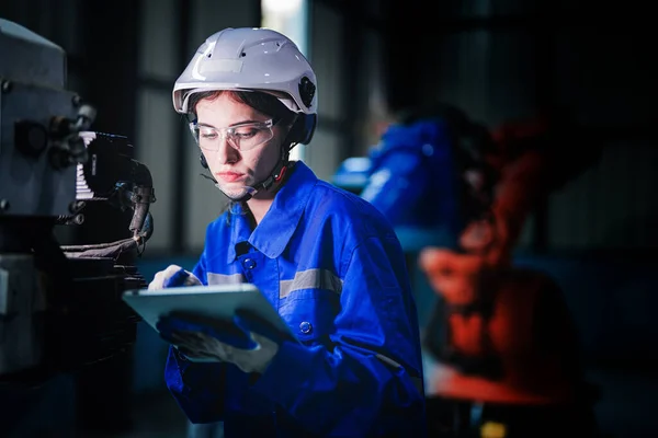 Factory engineer woman inspecting on machine with smart tablet. Worker works at machine robot arm. The welding machine with a remote system in an industrial factory. Artificial intelligence concept.