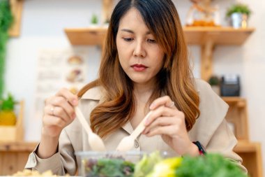 Genç Asyalı kadın salata sebzesiyle sağlıklı yiyecekler yiyor. Güzel bir iç mutfakta kilerde oturan bir kadın. Yerel ürünlerden ve malzemelerden alınan temiz diyet gıdaları taze pazardır..