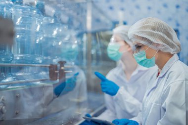 Bilim adamı, endüstriyel fabrikanın makine taşıma hattındaki su şişelerinin kalitesini kontrol ediyor. İçki üretim hattında veri kaydeden kadın işçi..